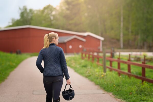 Kvinna i ridkläder går längs en grusväg och bär en ridhjälm i handen, på väg mot röda stallbyggnader. Röd trästaket och grönskande träd i bakgrunden, i mjukt kvällsljus.