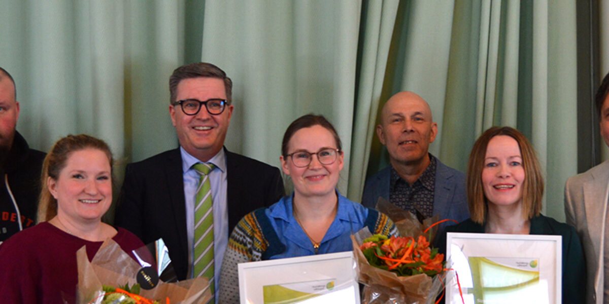 Sju personer står uppställda och ler mot kameran, några håller blombuketter och inramade diplom. De poserar tillsammans framför gröna gardiner i en inomhusmiljö, som vid en prisutdelning eller uppmärksammande.