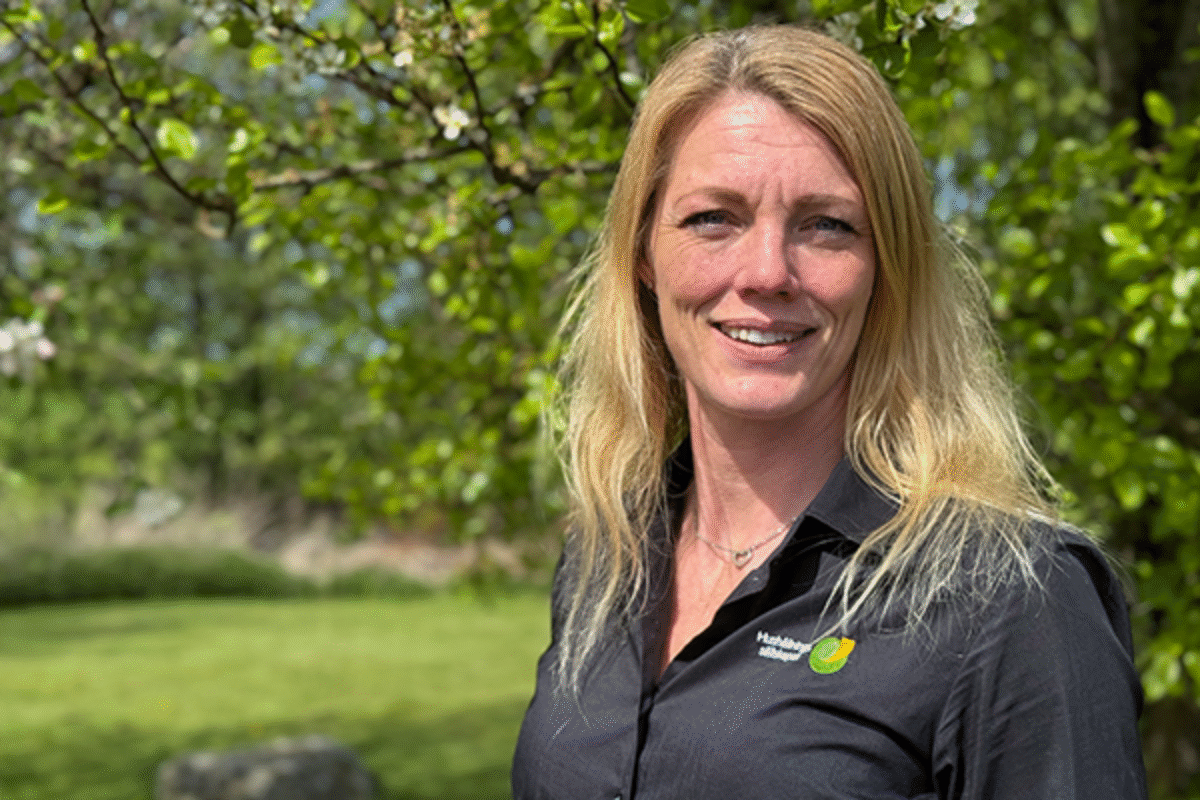Sara Löfman står utomhus framför ett blommande träd. Hon bär en svart skjorta med Hushållningssällskapets logotyp och ler mot kameran. Solen skiner och bakgrunden visar grönskande natur.