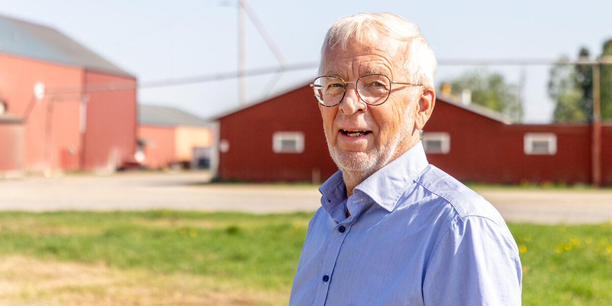 Äldre man med grått hår och glasögon står utomhus och tittar mot kameran. Han bär ljusblå skjorta och ler lätt, med röda ladugårdsbyggnader och gräsplan i bakgrunden i starkt dagsljus.