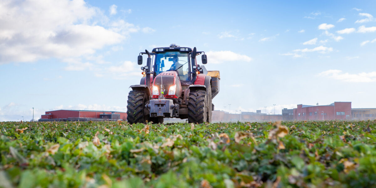 Röd traktor kör fram över en grön åker med en gödselspridare bakpå och damm som virvlar. Lågt fotovinkel med molnig blå himmel; industribyggnader syns i bakgrunden.
