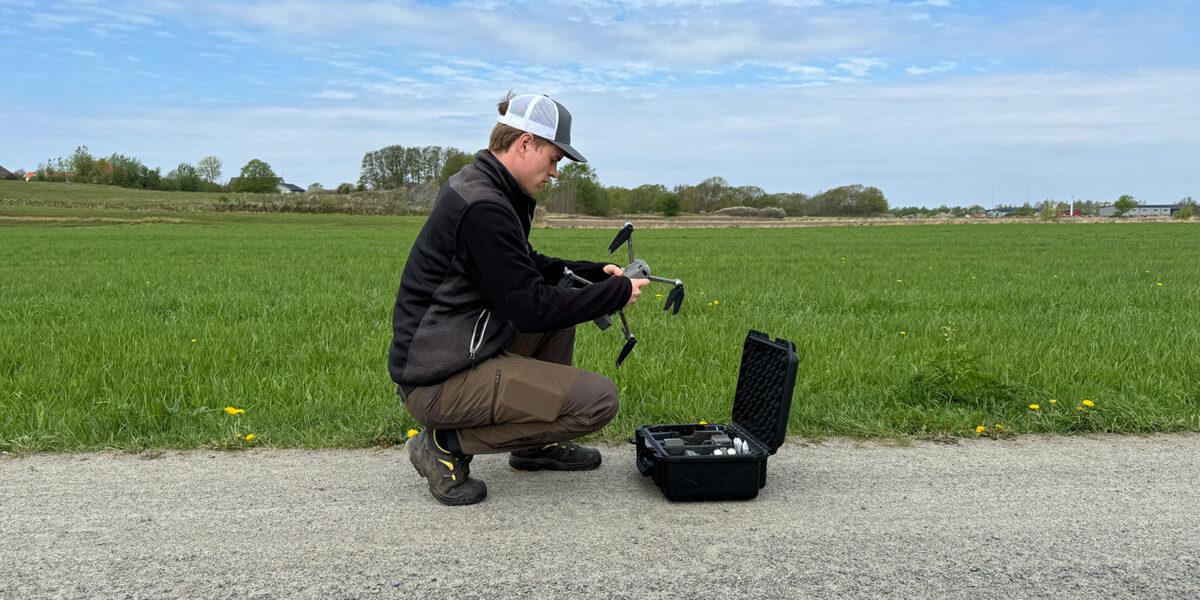 En person på huk monterar eller kontrollerar en drönare bredvid en öppen hård väska med utrustning. Scenen är utomhus på en grusväg intill en grön åker under blå himmel.