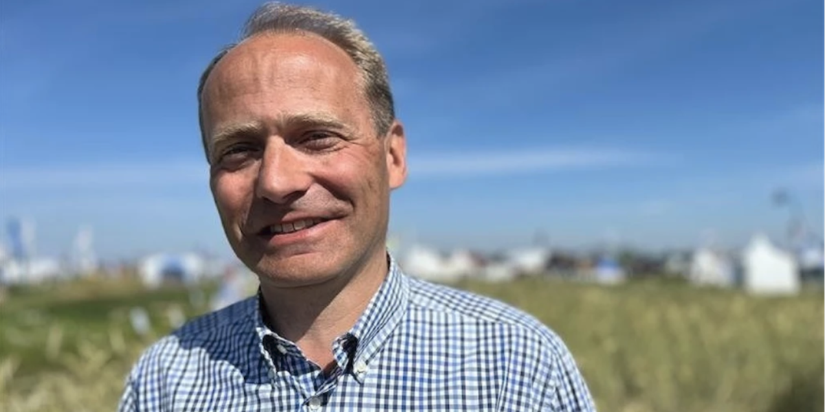 Leende man i blå- och vitrutiga skjorta står vänd mot kameran i solljus. Bakom honom syns en suddig äng och avlägsna vita tält eller byggnader under klarblå himmel.