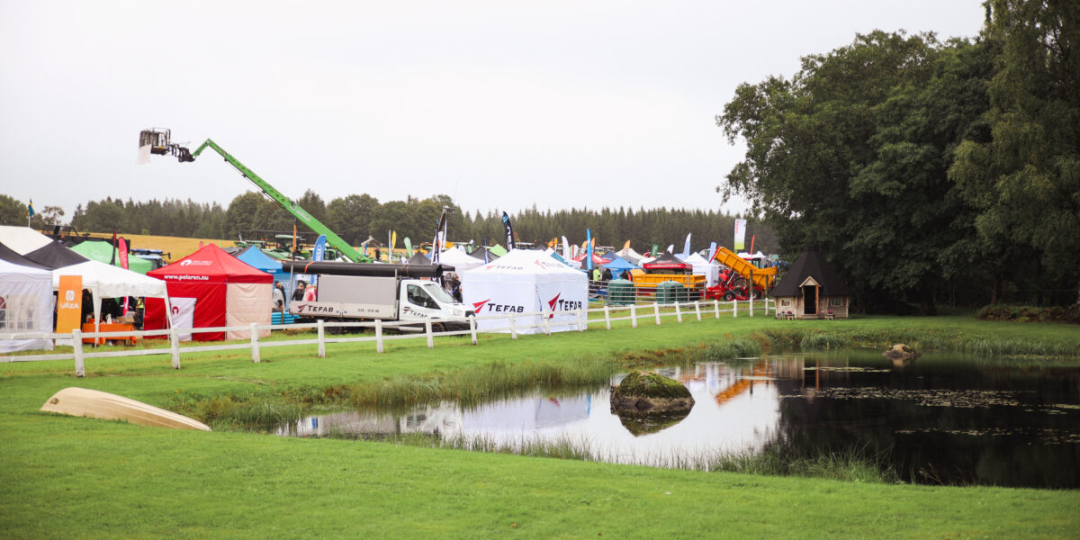 Utställningsområde med många tält och maskiner står uppställda längs ett vitt staket vid en liten damm. En grön skylift sträcker sig över mässan, och tält med texten “TEFAB” syns i bakgrunden.