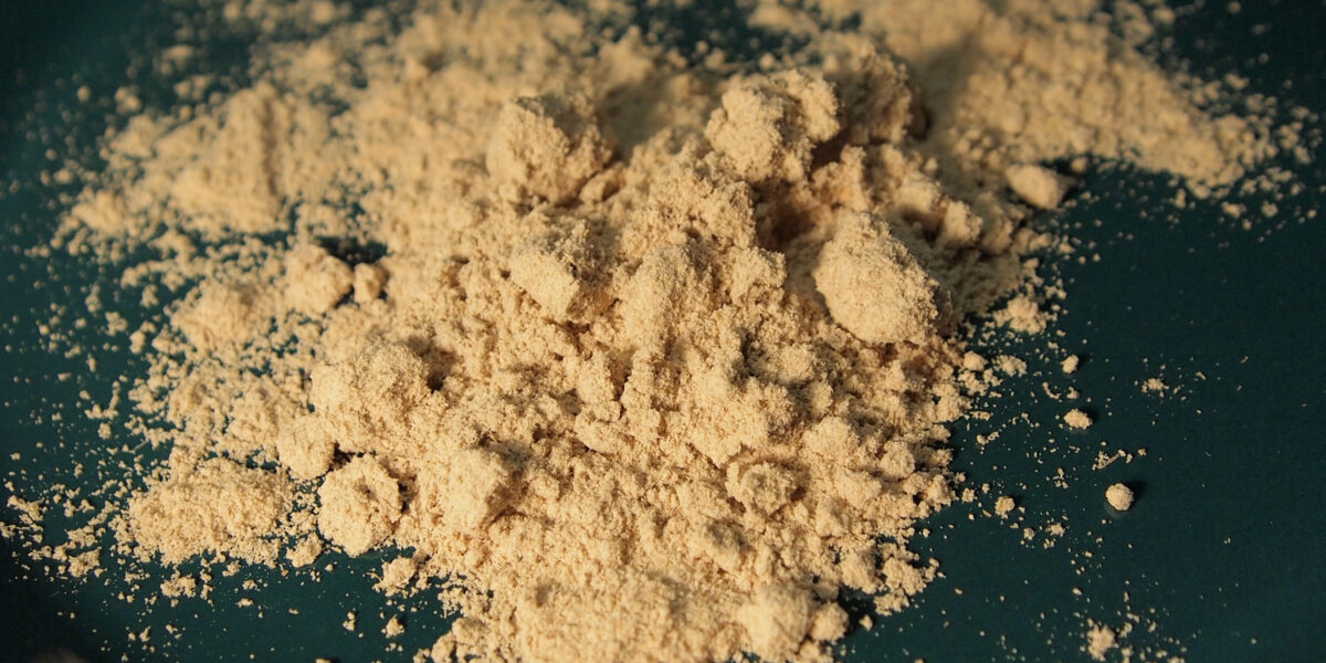 En hög ljusbeige mjöl eller pulver ligger i en liten hög och smulas i klumpar på en mörkgrön yta, med fint damm spritt runtom, som vid matlagning eller bakning.