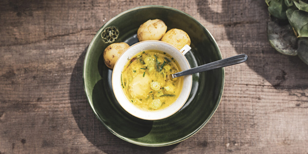 Skål med krämig gul soppa med örter och grönsaksbitar står på en grön tallrik, med sked och små bröd- eller potatisbitar bredvid. Allt ligger på ett rustikt träbord med gröna blad till höger.