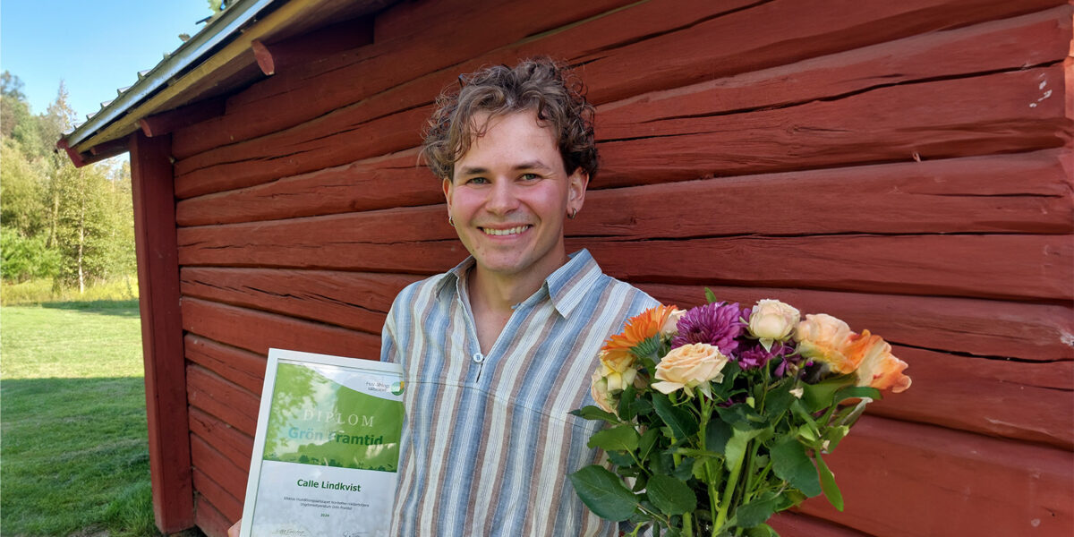 Leende person håller upp ett diplom och en bukett blommor framför en röd träbyggnad utomhus. Diplomet lyder: ”DIPLOM Grön framtid – Calle Lindkvist”. Grön gräsmatta och träd syns i bakgrunden.