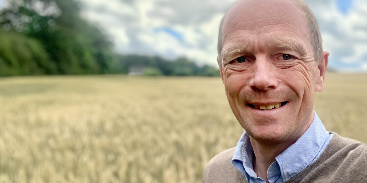 Leende, skallig man i brun tröja och ljusblå skjorta står och tittar mot kameran vid kanten av ett gyllene spannmålsfält, med suddig skog i bakgrunden och molnig blå himmel.