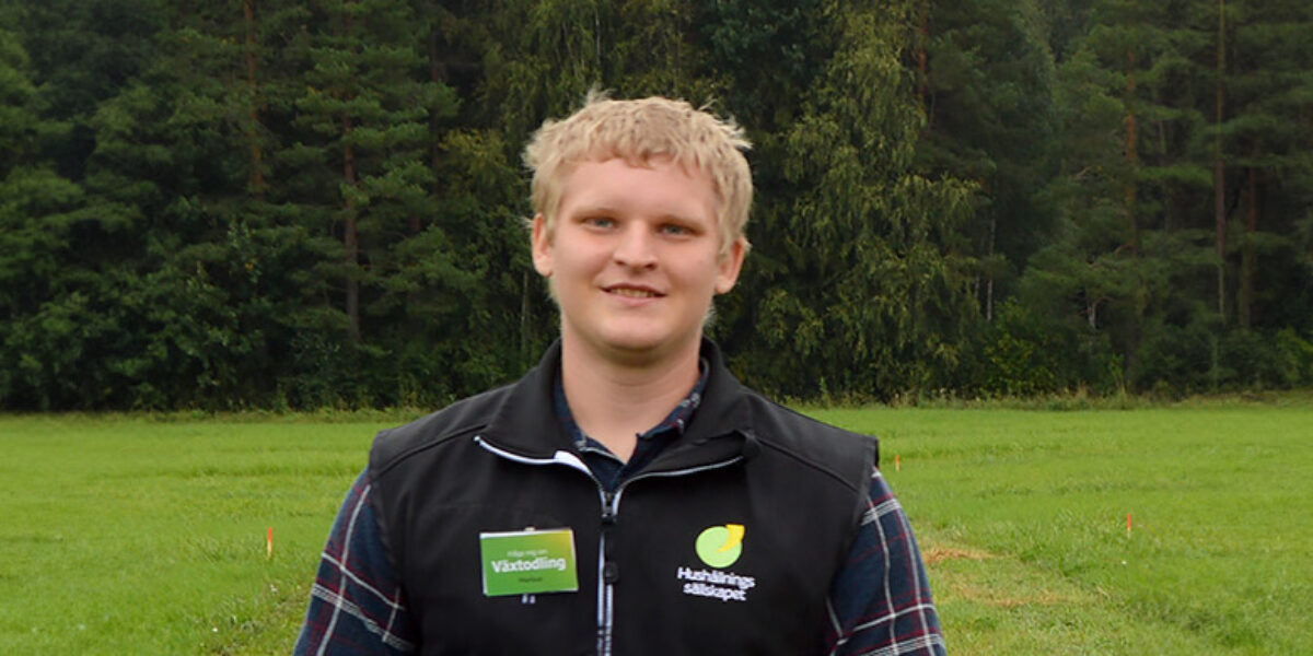 Ung man står och ler på en grön åker framför en skogskant. Han bär rutig skjorta och svart väst med logga samt namnbricka med texten ”Växtodling”, som antyder arbete inom lantbruk.