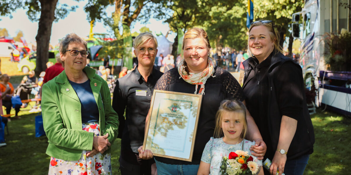 Fyra kvinnor poserar och ler utomhus på ett sommarmöte; en håller ett inramat diplom med texten ”Diplom”, och ett barn framför håller en blomsterbukett. I bakgrunden syns träd, folk och fordon.