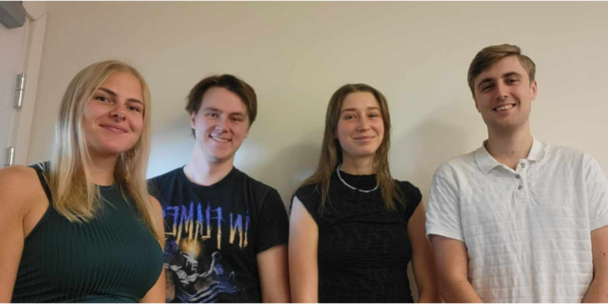 Fyra unga vuxna står sida vid sida och ler mot kameran, fotograferade inomhus mot en ljus vägg. De bär vardagskläder, bland annat en svart T-shirt med texten “IN FLAMES” och en vit pikétröja.