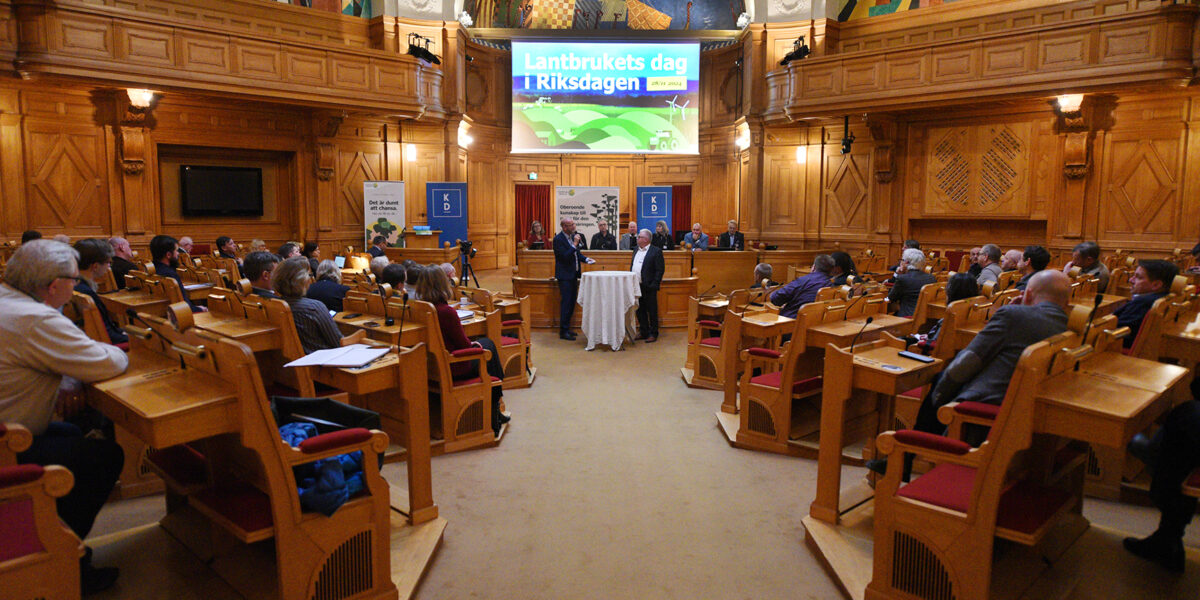 Åhörare sitter i träbänkar och lyssnar på talare vid ett runt bord längst fram i en rikt dekorerad plenisal. Stor skärm visar texten ”Lantbrukets dag i Riksdagen” över gröna fält.