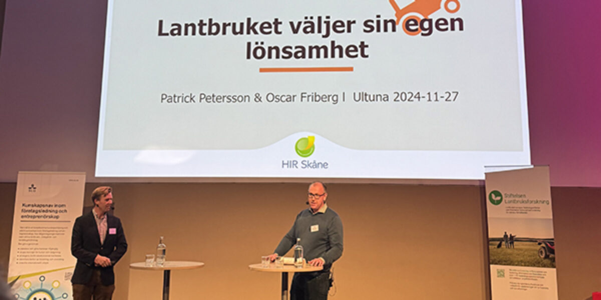 Två föreläsare står på en scen och presenterar en jordbruksekonomisk konferens. På storbildsskärmen visas texten “Lantbruket väljer sin egen lönsamhet”, “Patrick Petersson & Oscar Friberg | Ultuna 2024-11-27” och “HIR Skåne”.