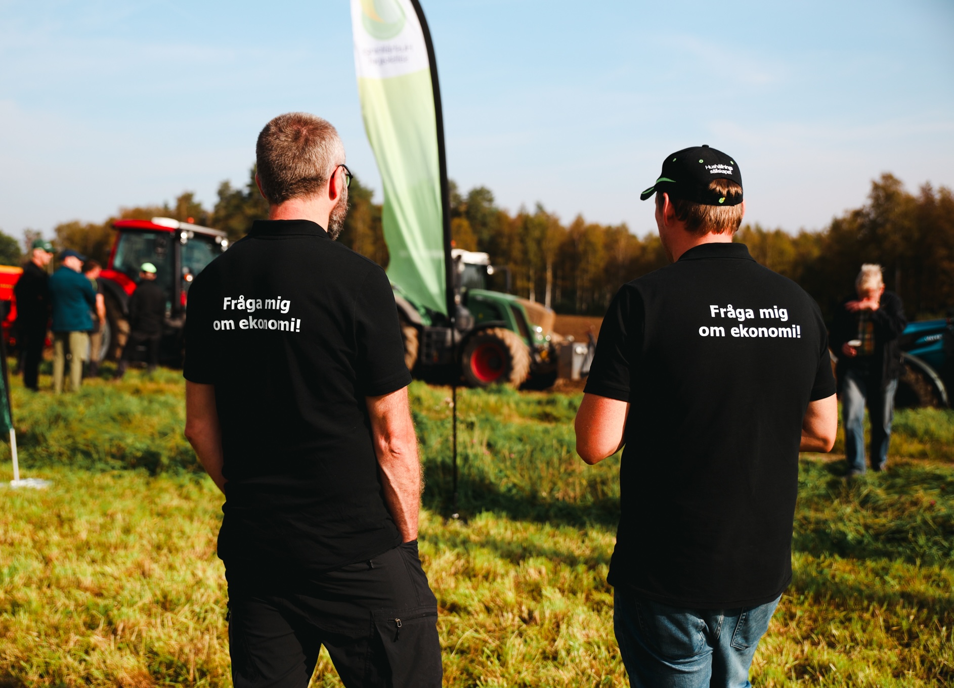 Två män i svarta pikétröjor står med ryggen mot kameran och pratar på en lantbruksmässa. Text på tröjorna: ”Fråga mig om ekonomi!”. I bakgrunden syns traktorer, besökare och en grön flagga.