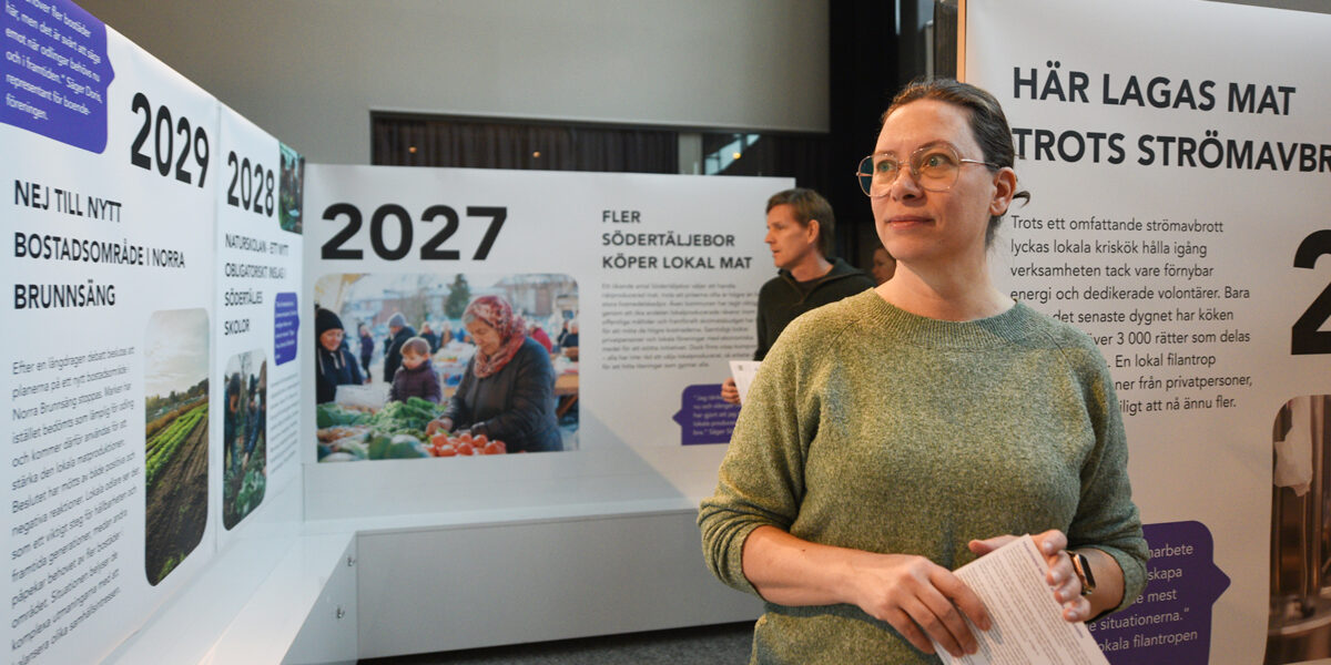Besökare med glasögon håller ett informationsblad och tittar åt sidan i en utställningslokal, framför väggpaneler med årtal 2027–2029 och rubrikerna ”FLER SÖDERTÄLJEBOR KÖPER LOKAL MAT” samt ”HÄR LAGAS MAT TROTS STRÖMAVBROTT”.