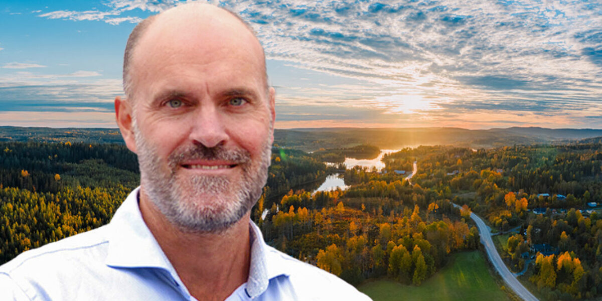 Leende skallig man med skägg i vit skjorta blickar mot kameran, framför en vid utsikt över skog och sjöar i solnedgång; en väg slingrar sig genom höstfärgade träd under molnig himmel.