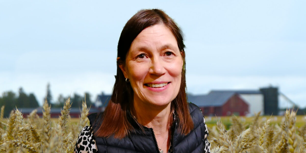 Leende kvinna står i en mogen vetåker och tittar mot kameran, klädd i svart vadderad väst och mönstrad tröja. I bakgrunden syns gårdsbyggnader och träd under en ljus, molnig himmel.