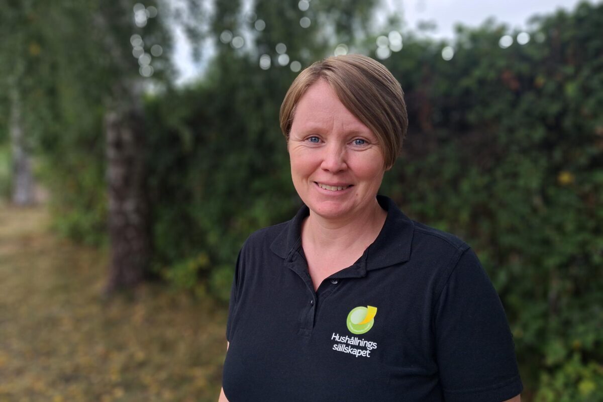 Leende person med kort brunt hår tittar mot kameran och står utomhus framför suddiga träd och buskar. Bär svart pikétröja med logga och texten “Hushållnings sällskapet” på bröstet.