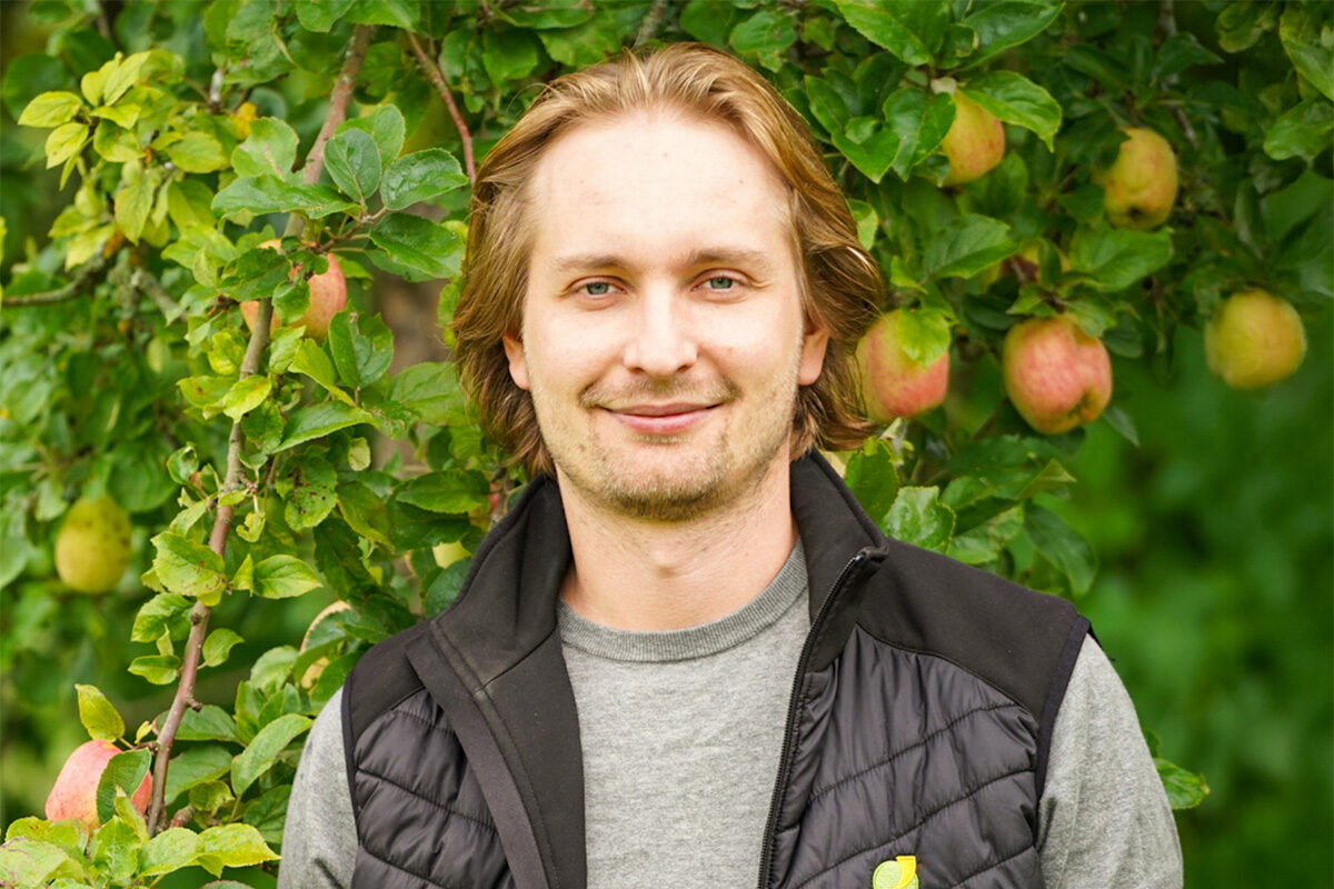 Leende man med halvlångt hår står och tittar mot kameran, iklädd grå tröja och svart vadderad väst. Bakom honom syns ett frodigt äppelträd med gröna blad och flera röda äpplen.