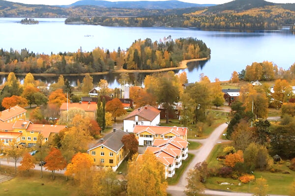 Flygfoto visar ett lantligt bostadsområde med flera gula och vita hus med röda tak, omgivna av höstfärgade träd och grusvägar. I bakgrunden syns en stor sjö med skogsklädda halvöar och kullar.
