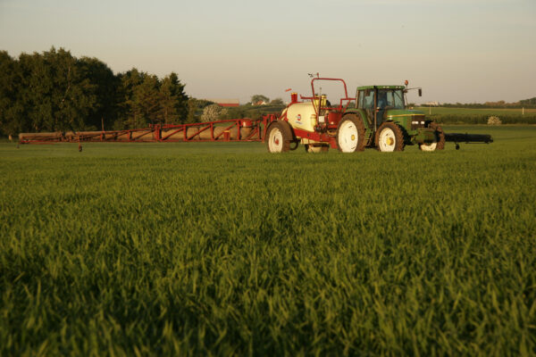 Grön traktor med stor röd fältspruta kör över en grön åker och sprider växtskydd eller gödning. Långa sprutbommar är utfällda åt sidan, med träd och öppet jordbrukslandskap i bakgrunden.