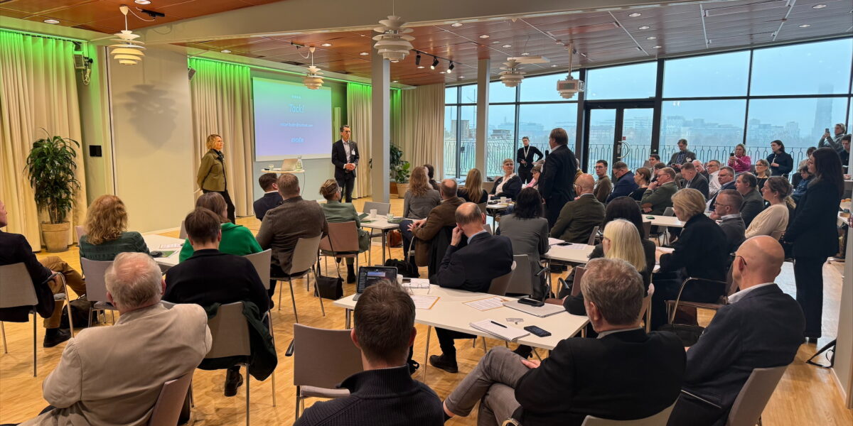 Deltagare sitter vid bord och lyssnar när två talare står längst fram i en konferenssal; en person ställer en fråga. Projektorduken visar texten “Tack!”. Stora fönster och grön belysning ramar in lokalen.