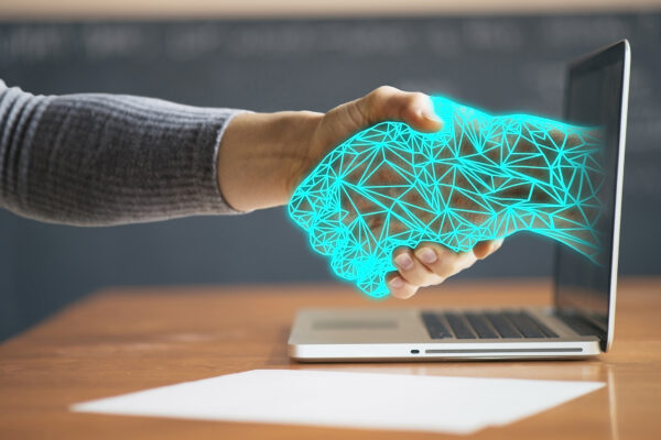 En mänsklig hand skakar hand med en neonblå, polygonformad digital hand som kommer ut ur en laptopskärm, på ett skrivbord. Motivet symboliserar säker digital interaktion, förtroende och cybersäkerhet.
