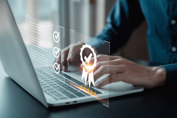 Händer på en person som skriver på en bärbar dator, medan en transparent digital panel visar checklista med bockar, ett certifieringsmärke med bock och fem stjärnor som antyder kvalitetsgranskning eller godkännande.