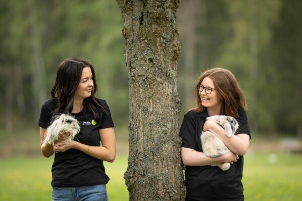 Två kvinnor i svarta tröjor håller varsitt smådjur och ler mot varandra utomhus. Den ena bär ett långhårigt marsvin, den andra en kanin, med en trädstam mellan dem i en grön parkmiljö.