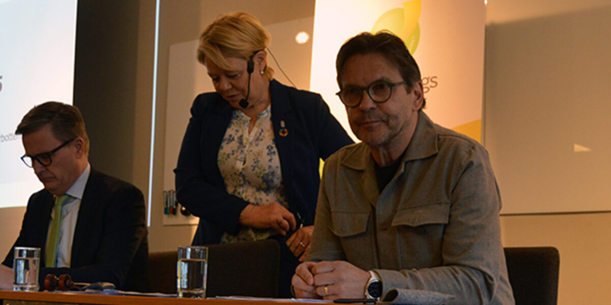 Tre personer leder ett möte vid ett bord; en man skriver anteckningar, en kvinna står bakom och förbereder sig, och en man sitter vänd mot publiken. Projektor visar: “Årsmöte 2025”.