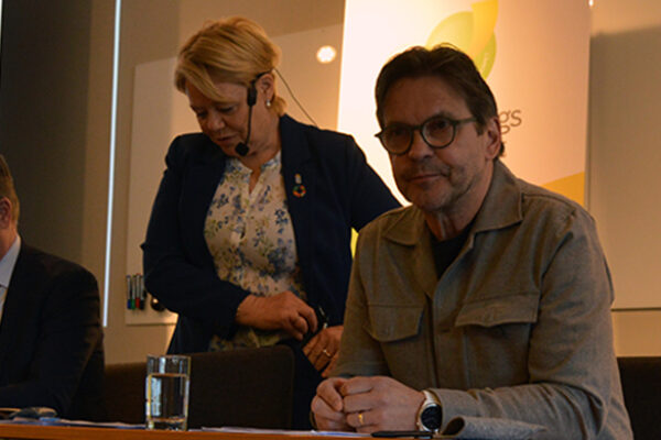 Tre personer leder ett möte vid ett bord; en man skriver anteckningar, en kvinna står bakom och förbereder sig, och en man sitter vänd mot publiken. Projektor visar: “Årsmöte 2025”.