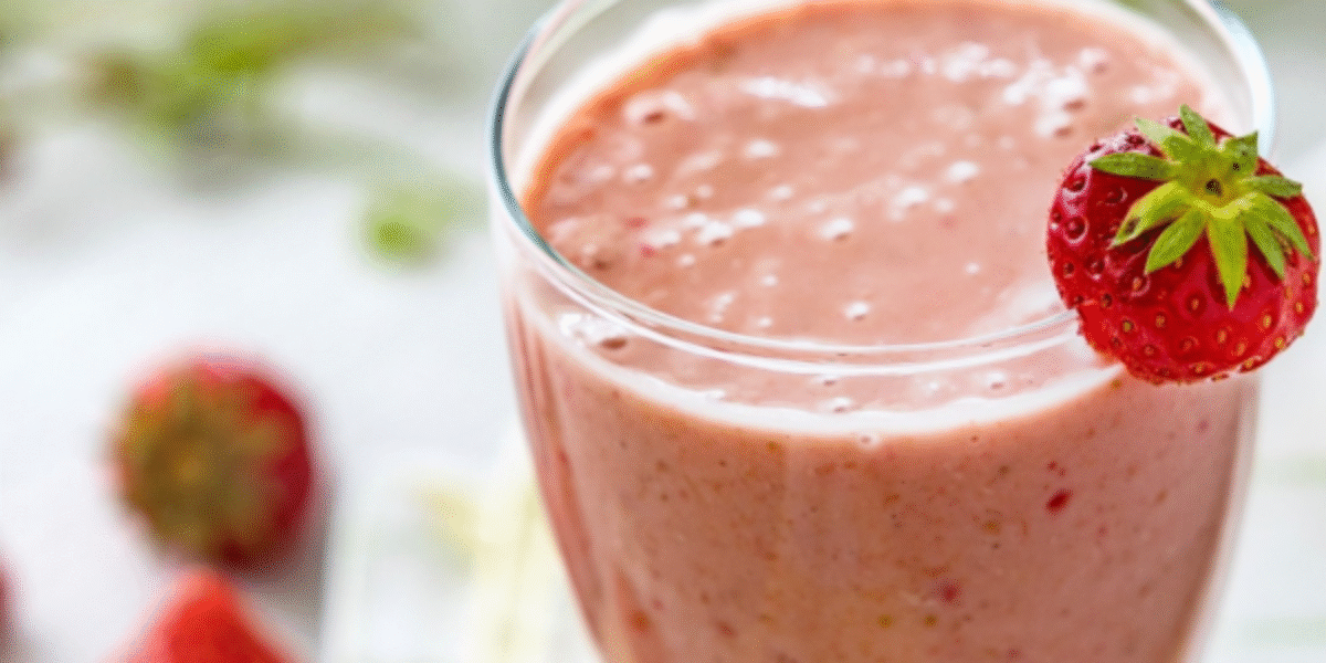 Ett glas med ljusrosa jordgubbssmoothie står i närbild, med en färsk jordgubbe som dekoration på kanten. I suddig bakgrund syns fler jordgubbar och grönska på en ljus yta.