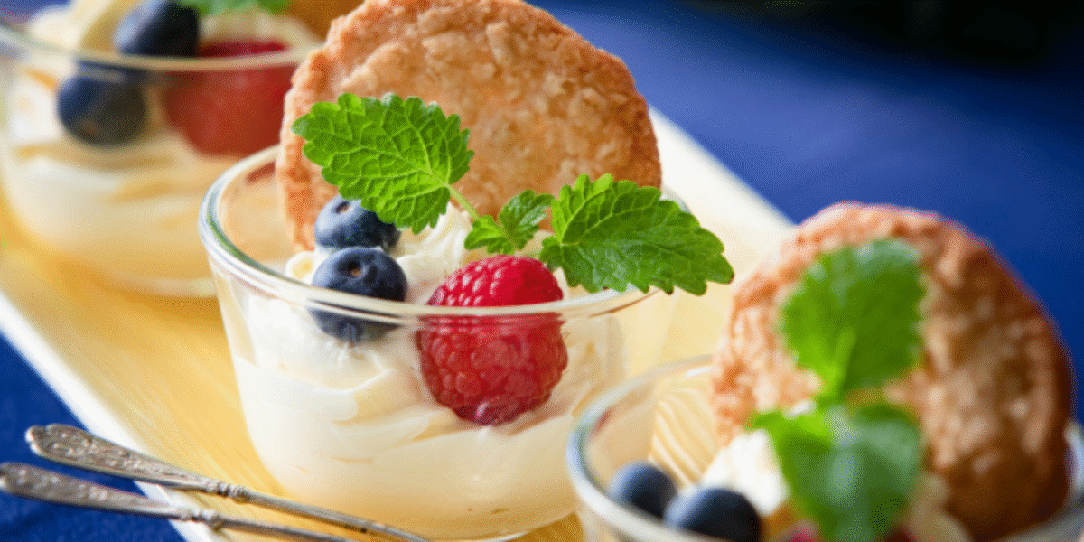 Tre glasdesserter med krämig vanilj, toppade med hallon, blåbär och myntablad, står på ett avlångt fat. En rund småkaka sticker upp i varje glas, med skedarna placerade intill på blå duk.