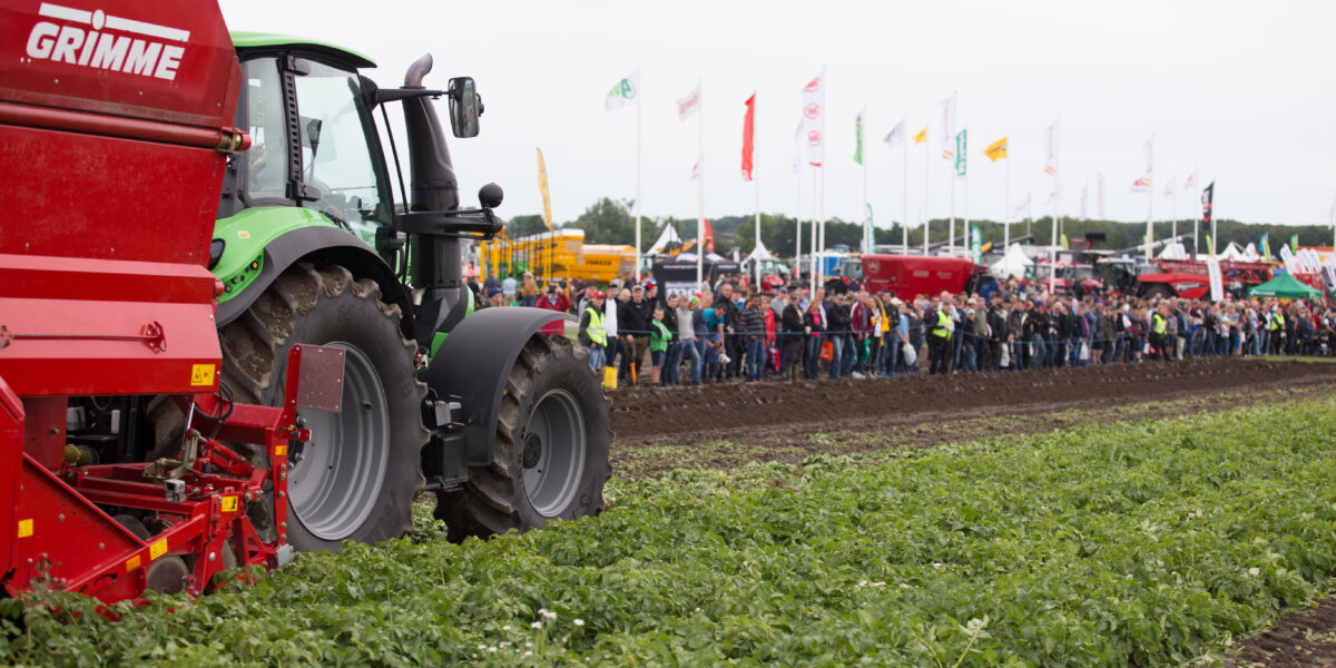 Grön traktor som drar en röd GRIMME-maskin kör längs en potatisåker under en maskindemonstration. En stor publik står bakom avspärrning och tittar, med mässflaggor och utställningsfordon i bakgrunden.