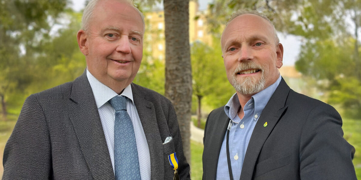 Två män i kostym står sida vid sida och ler mot kameran. Den ena bär blå slips och en medalj i blågult band, den andra har skägg, blå skjorta och namnbricka, utomhus bland träd.