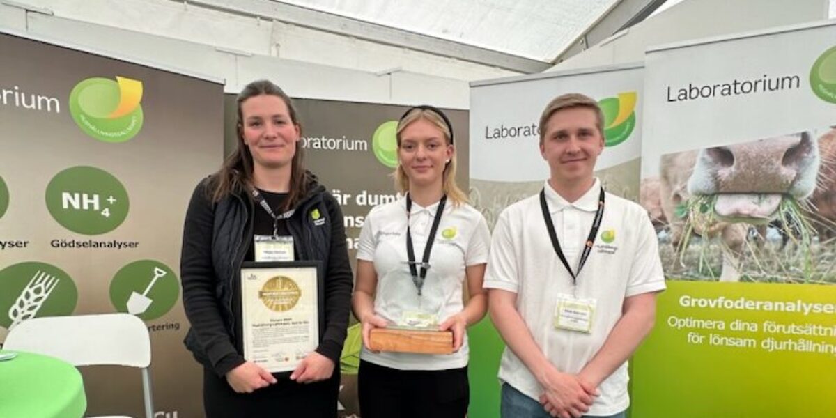 Tre personer på en mässa står framför rollups för ”Laboratorium”; två visar upp en inramad utmärkelse och en glasplakett. Bakgrunden har texten ”Grovfoderanalyser” och ”Optimera dina förutsättningar för lönsam djurhållning”.
