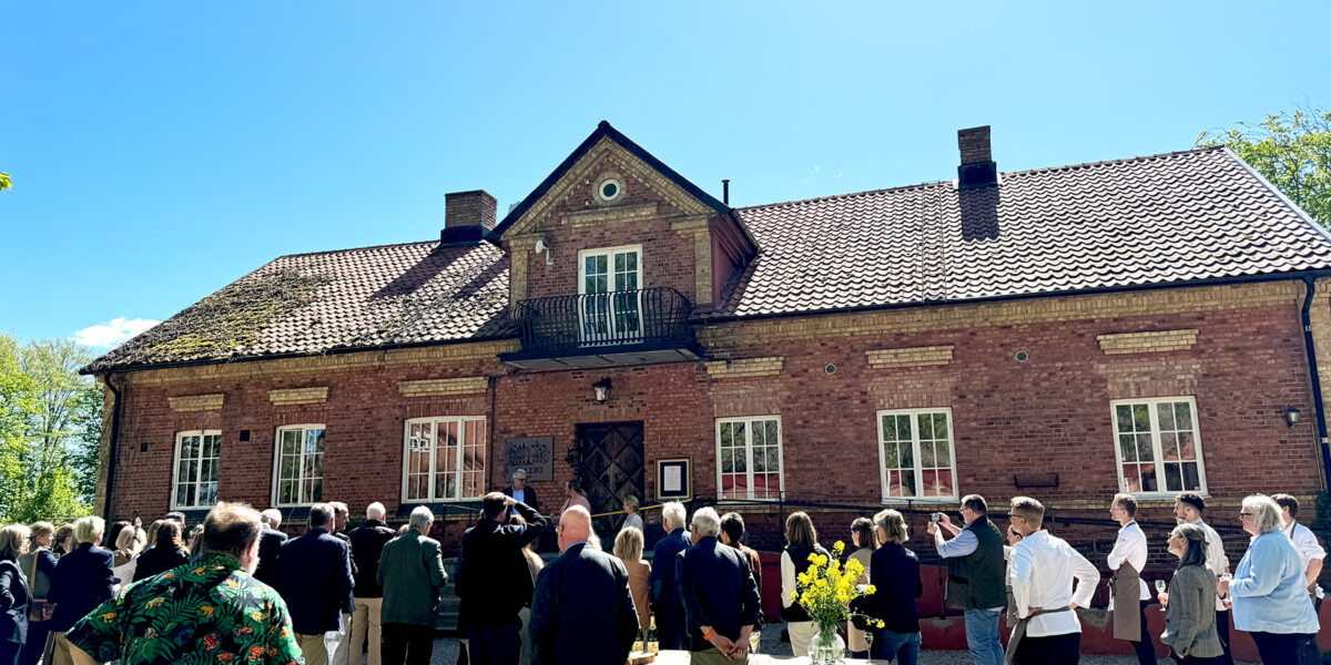 En grupp människor samlas och lyssnar utanför en stor tegelbyggnad med balkong, som vid en invigning. Några fotograferar och flera står runt ett bord med gul blomsterbukett i solsken, mot klarblå himmel.