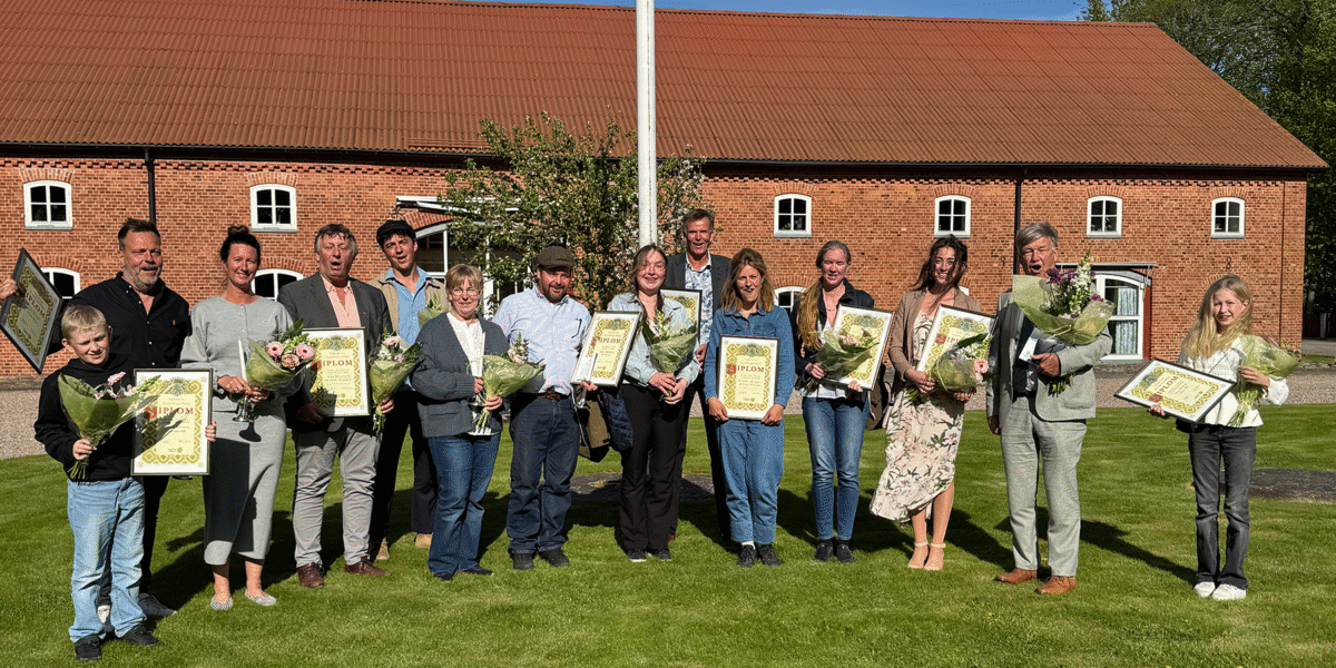 Grupp pristagare står på en gräsmatta och poserar med buketter och inramade diplom framför en röd tegelbyggnad med brunt tak och flaggstång; diplomen har rubriken “DIPLOM” och flera ler mot kameran.