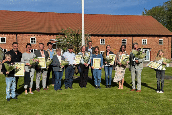 Grupp pristagare står på en gräsmatta och poserar med buketter och inramade diplom framför en röd tegelbyggnad med brunt tak och flaggstång; diplomen har rubriken “DIPLOM” och flera ler mot kameran.