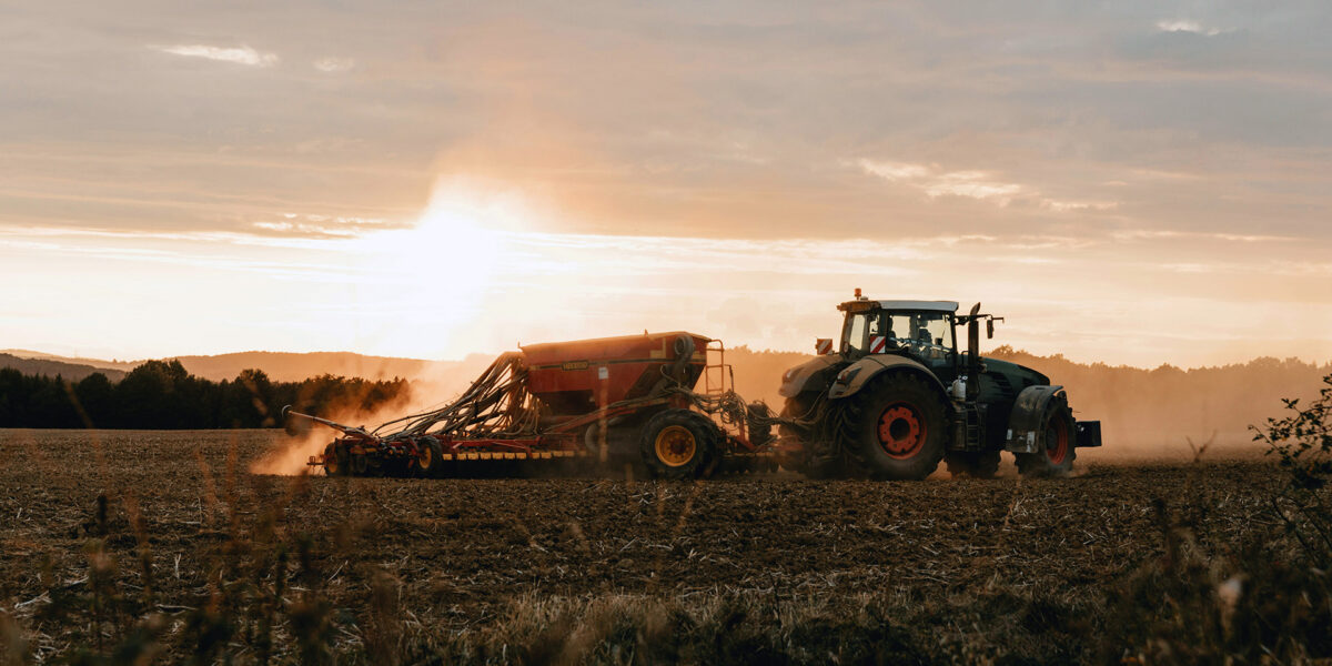 Traktor drar en såmaskin över en nyplöjd åker och virvlar upp damm. Lågt kvällsljus och solnedgång i bakgrunden ger siluett mot himlen, med skog och mjuka kullar vid horisonten.