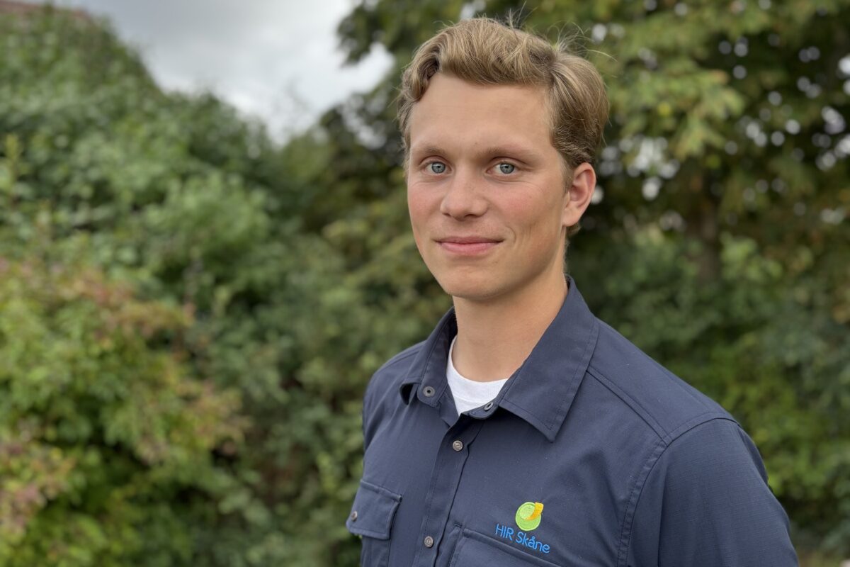 Ung man står utomhus och tittar mot kameran med ett svagt leende, iklädd mörkblå skjorta med broderad logotyp och texten “HIR Skåne”. Grönskande träd och buskar i bakgrunden under molnig himmel.