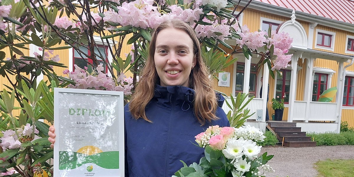 Leende kvinna håller upp ett diplom och en bukett med rosa och vita blommor framför ett gult hus med röd takplåt och veranda, omgiven av blommande buskar. Text på diplomet: “DIPLOM Linnea Hedborg”.