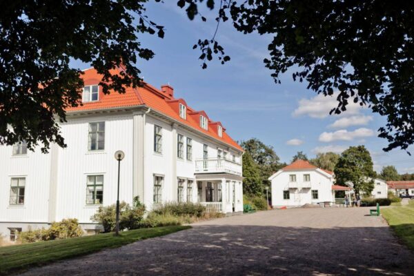 Vit träbyggnad med rött tegeltak står längs en grusad uppfart, delvis inramad av trädgrenar, med veranda och många fönster. I bakgrunden syns en mindre liknande byggnad, bänkar och några personer vid gårdsplanen.