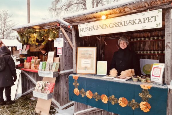 Person står och säljer varor i en vinterpyntad marknadsbod med skylten ”HUSHÅLLNINGSSÄLLSKAPET”. På disken syns burkar och broschyrer samt en QR-skylt med ”50 kr” och numret ”123 651 62 56”, snö runtom.