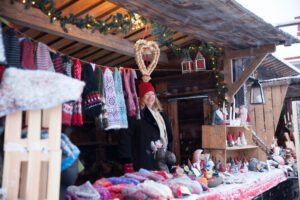Kvinna i röd mössa står och ler i en julmarknadsbod medan snö faller. Runt henne hänger stickade vantar och halsdukar, och på bordet ligger garn och små tomtar; granris och ljusslingor pryder taket.