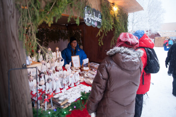 Besökare i vinterjackor handlar vid en julmarknadsbod där säljaren står bakom ett bord med handgjorda änglar och små dekorationer. Granris pryder taket och en skylt ovanför säger “Pysselboden” i snöig vintermiljö.