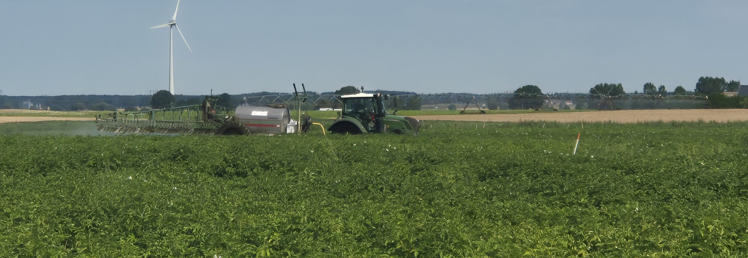 Traktor med tankvagn sprider vätska över en grön odling på ett fält, medan dimma av sprutmedel syns bakom redskapet. I bakgrunden syns en stor vindkraftverk och bevattningsanläggning under klar himmel.