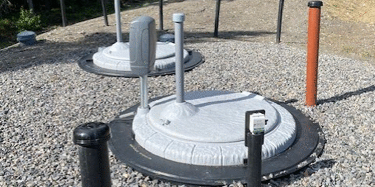 Två runda, grå lock med uppstickande rör och ett elskåp står på en grusad yta, omgivna av flera stolpar och skyddspinnar. Installationerna ser ut som tekniska brunnar för vatten eller avlopp utomhus.