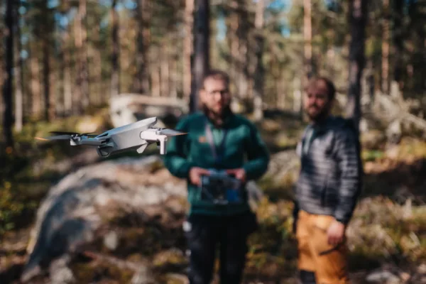 En grå quadcopter-drönare svävar i förgrunden medan två män står suddigt i bakgrunden i en skog; den ena håller en fjärrkontroll, vilket visar att de flyger och övervakar drönaren utomhus.