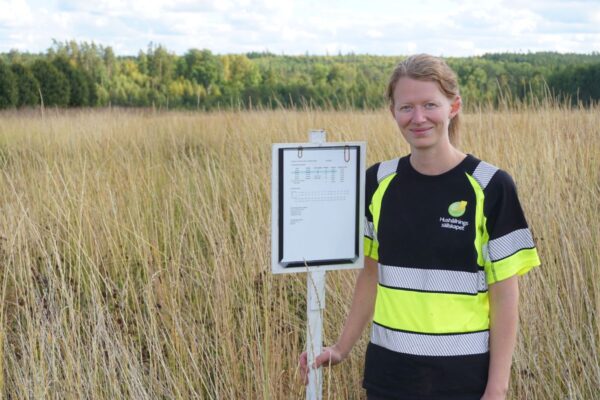 Kvinna i gul reflexväst står i ett högt gräsfält och håller i en vit stolpe med en inramad pappersskylt med tabell. Hon ler mot kameran, med skog och molnig himmel i bakgrunden.